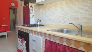 A kitchen with a gas stove, sink, and tiled backsplash. There are cabinets and a clock on the wall. - Monolocale Rosso (Turin)