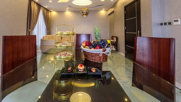 A modern hotel lobby with a glass table displaying various desserts, a wicker fruit basket, and a chandelier. - Arabella Residence (New Cairo)