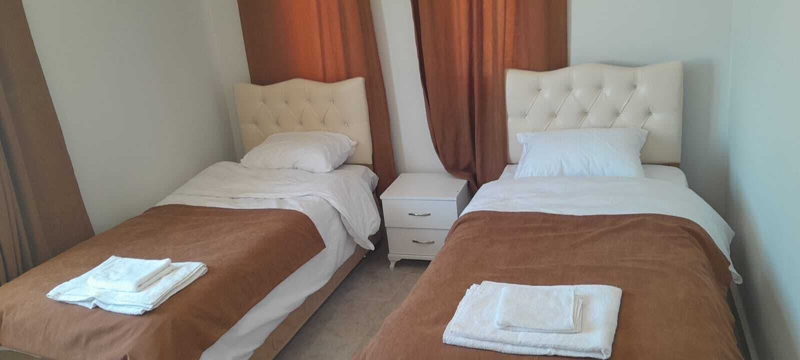 A hotel room with two beds, each with a brown bedspread and white pillows. There are brown curtains on the windows, and a white nightstand between the beds.