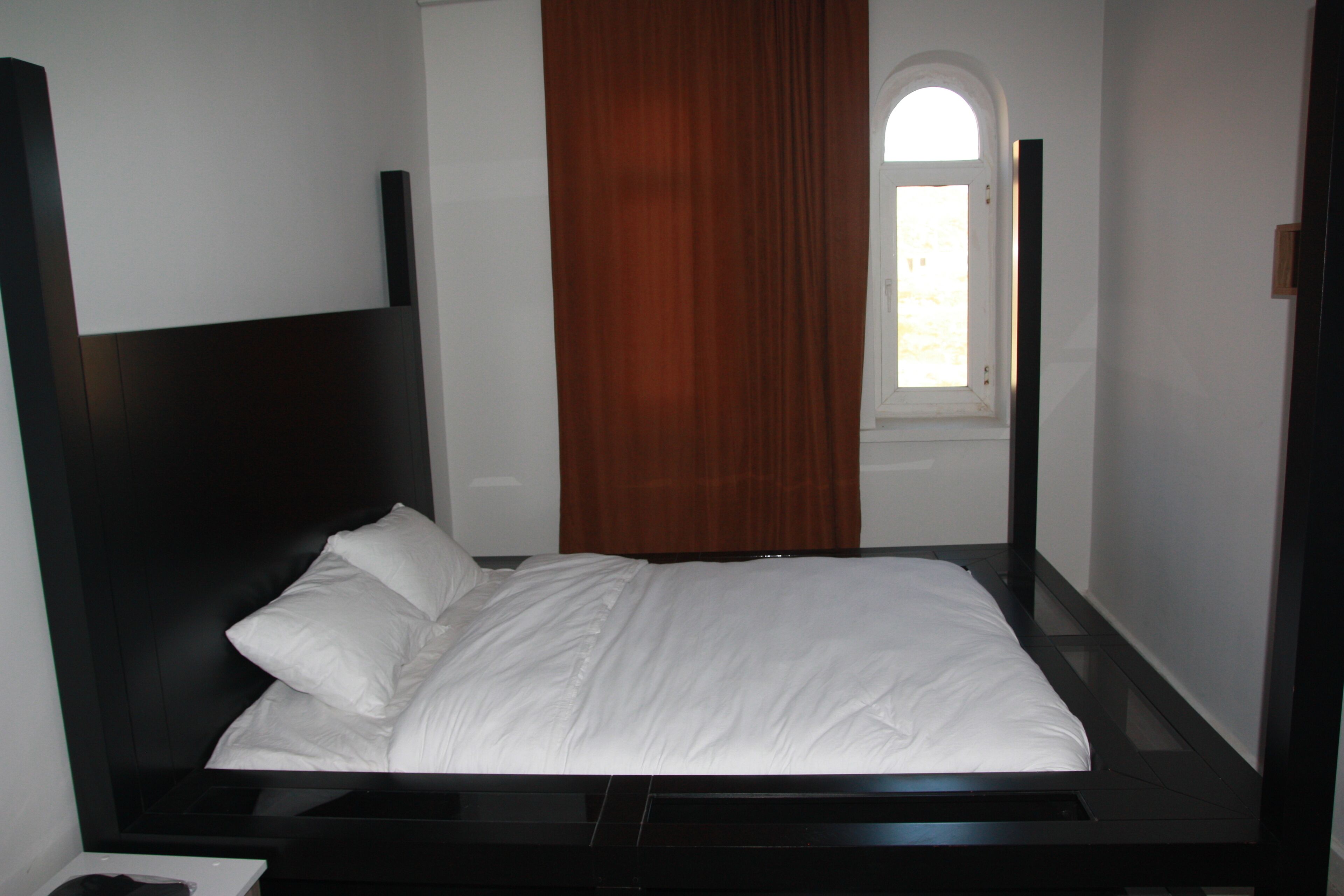Una cama bien hecha con ropa blanca en una habitación con cabecera oscura y ventana con cortina marrón.