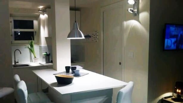 Una cocina moderna con una isla blanca, dos taburetes de bar, una lámpara colgante y un televisor montado en la pared.