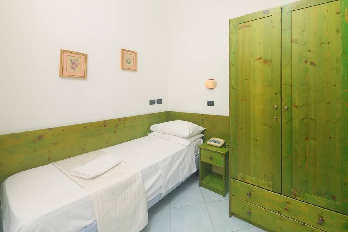 A single bed with a white mattress and a green headboard, a small bedside table with a lamp, and a green wardrobe with two doors.