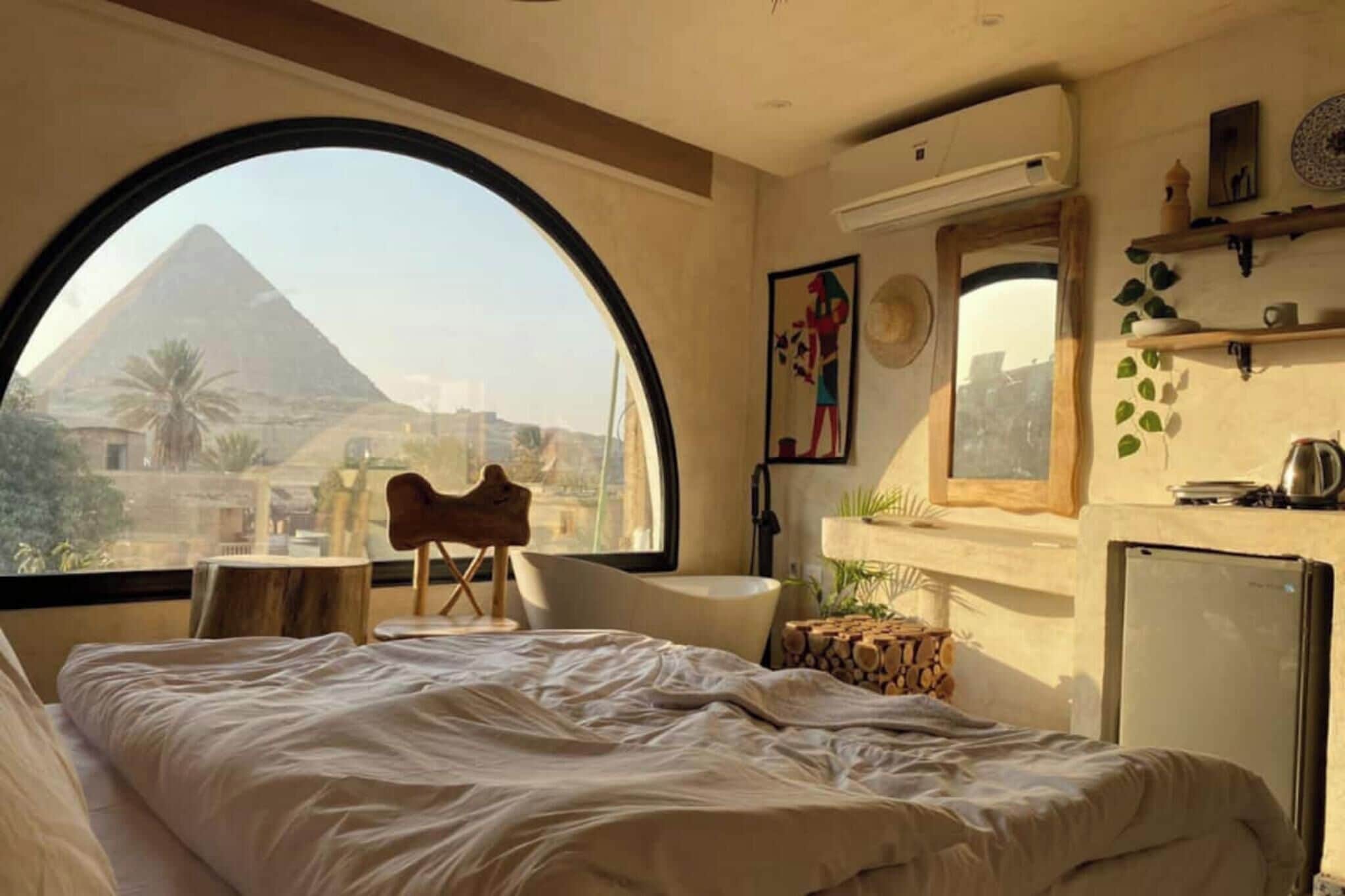 Habitación de hotel con un amplio ventanal arqueado, una cama, una mesita, una silla y un refrigerador.