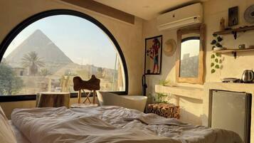 Habitación de hotel con un amplio ventanal arqueado, una cama, una mesita, una silla y un refrigerador.