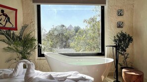 Un baño con una bañera independiente, un ventanal grande y vistas a un paisaje tropical.