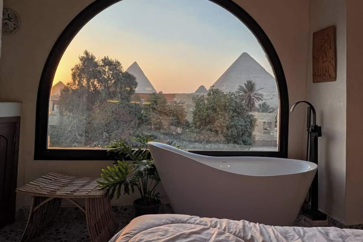 A room with a large arched window, a freestanding bathtub, a bed, and a small table with plants.
