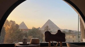 Une vue sur les pyramides, encadrée par une grande fenêtre en arc de cercle, une chaise en bois et une petite table sur laquelle se trouvent une tasse et un panier.