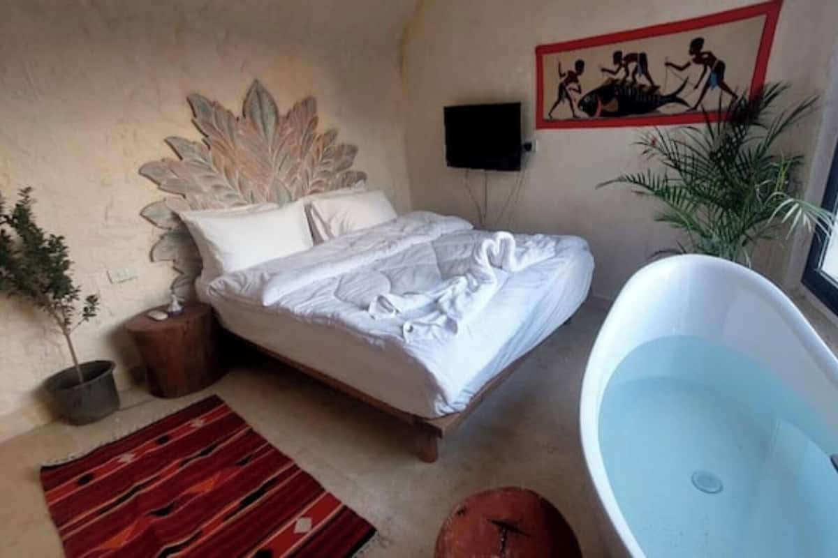 A bedroom with a bed, a large window, a bathtub, and a wall-mounted TV.