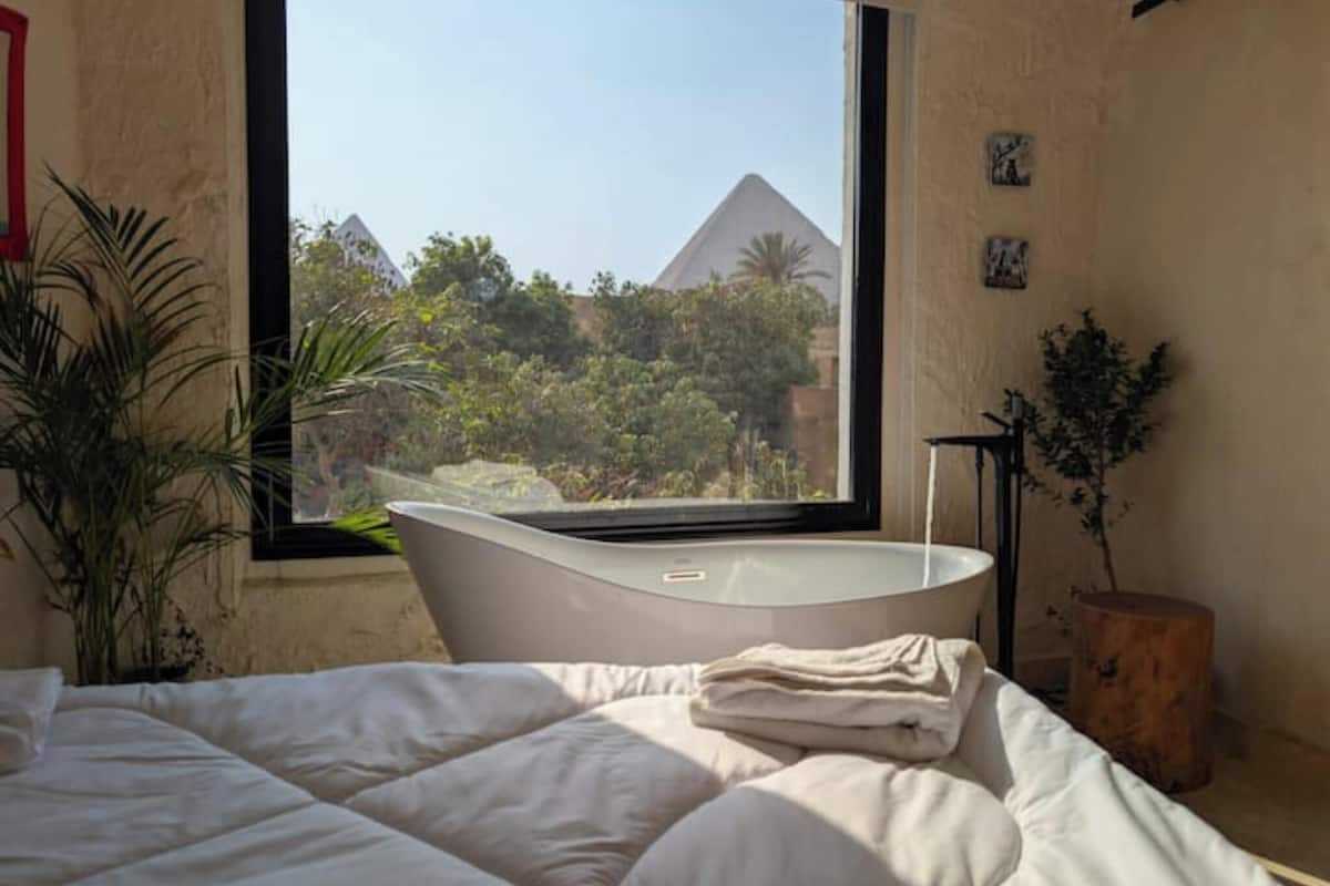 A bedroom with a large window, a bathtub, a bed, and a potted plant.