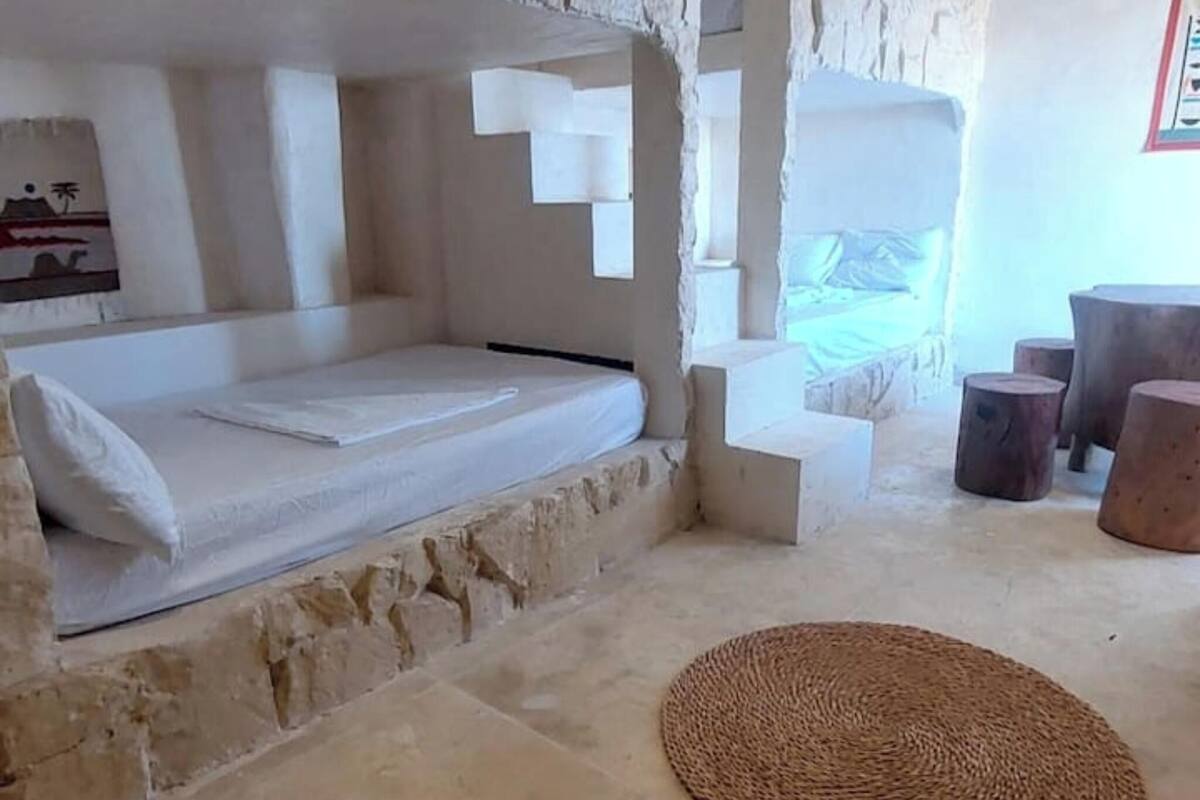 A stone-walled room with a bed, a woven rug, and wooden stools.