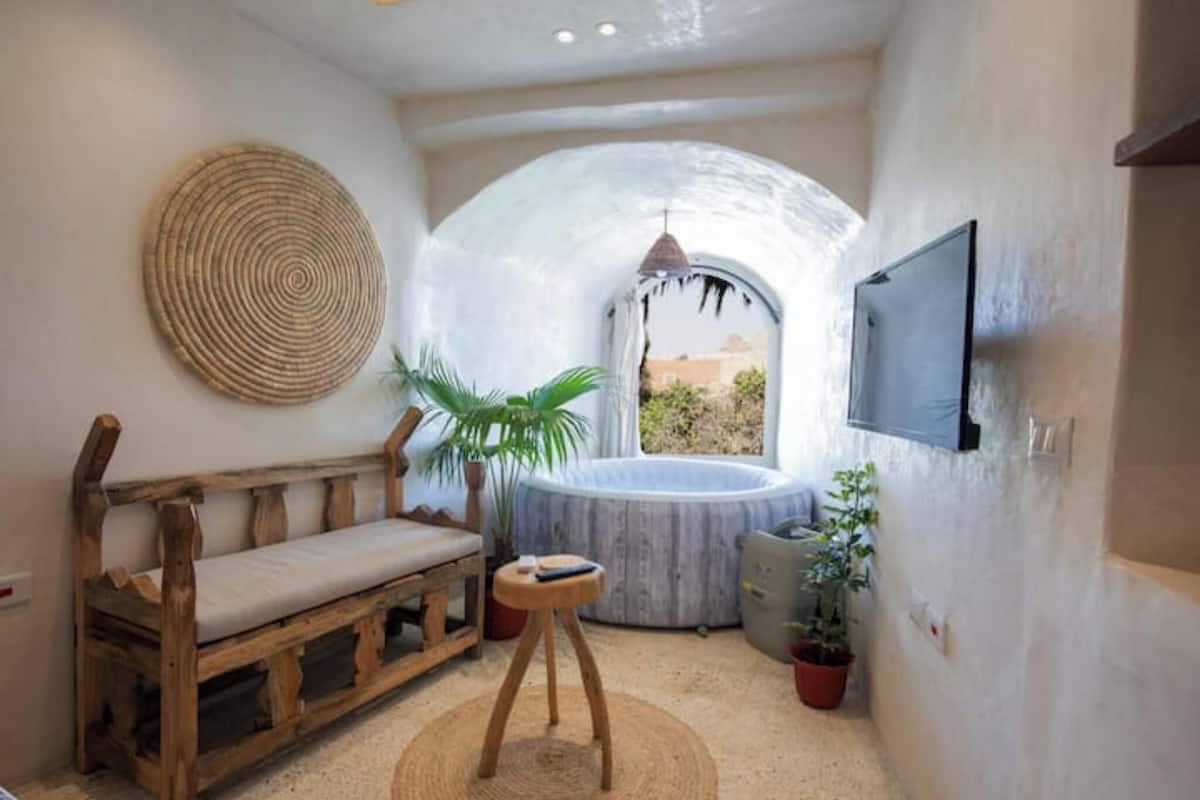 A cozy room with a wooden bench, a round woven rug, a small stool, a potted plant, a hot tub, and a TV mounted on the wall.