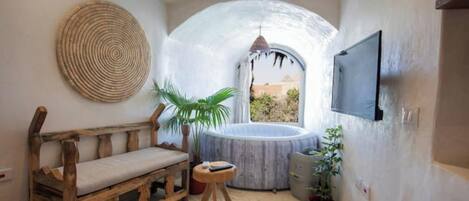 Una habitación acogedora con un banco de madera, un tapete redondo tejido, un taburete pequeño, una planta en maceta, un jacuzzi y un televisor montado en la pared.