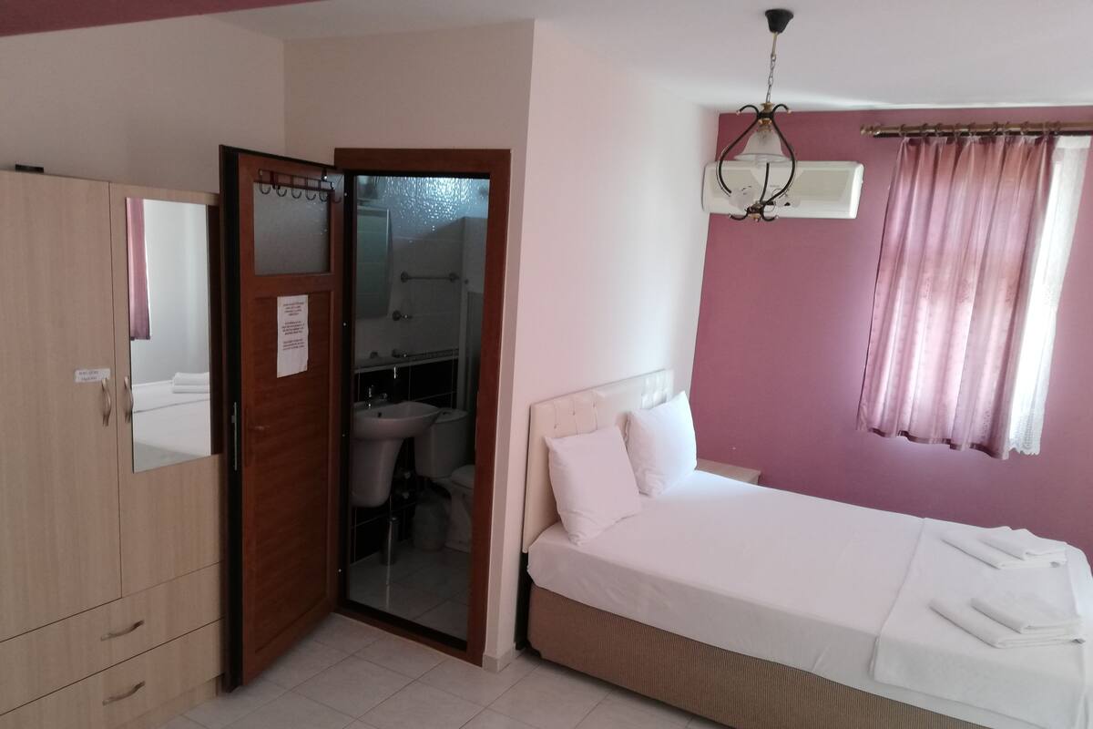 A bedroom with a bed, a wardrobe, a bathroom visible through an open door, and a window with curtains.