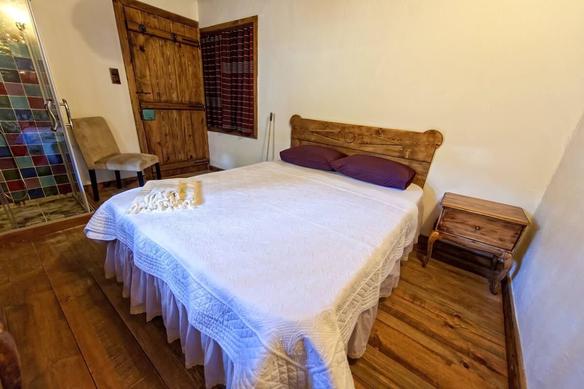 Un dormitorio con una cama de madera, una mesita de noche pequeña de madera, una silla y un vidrio que da a una zona con baldosas.