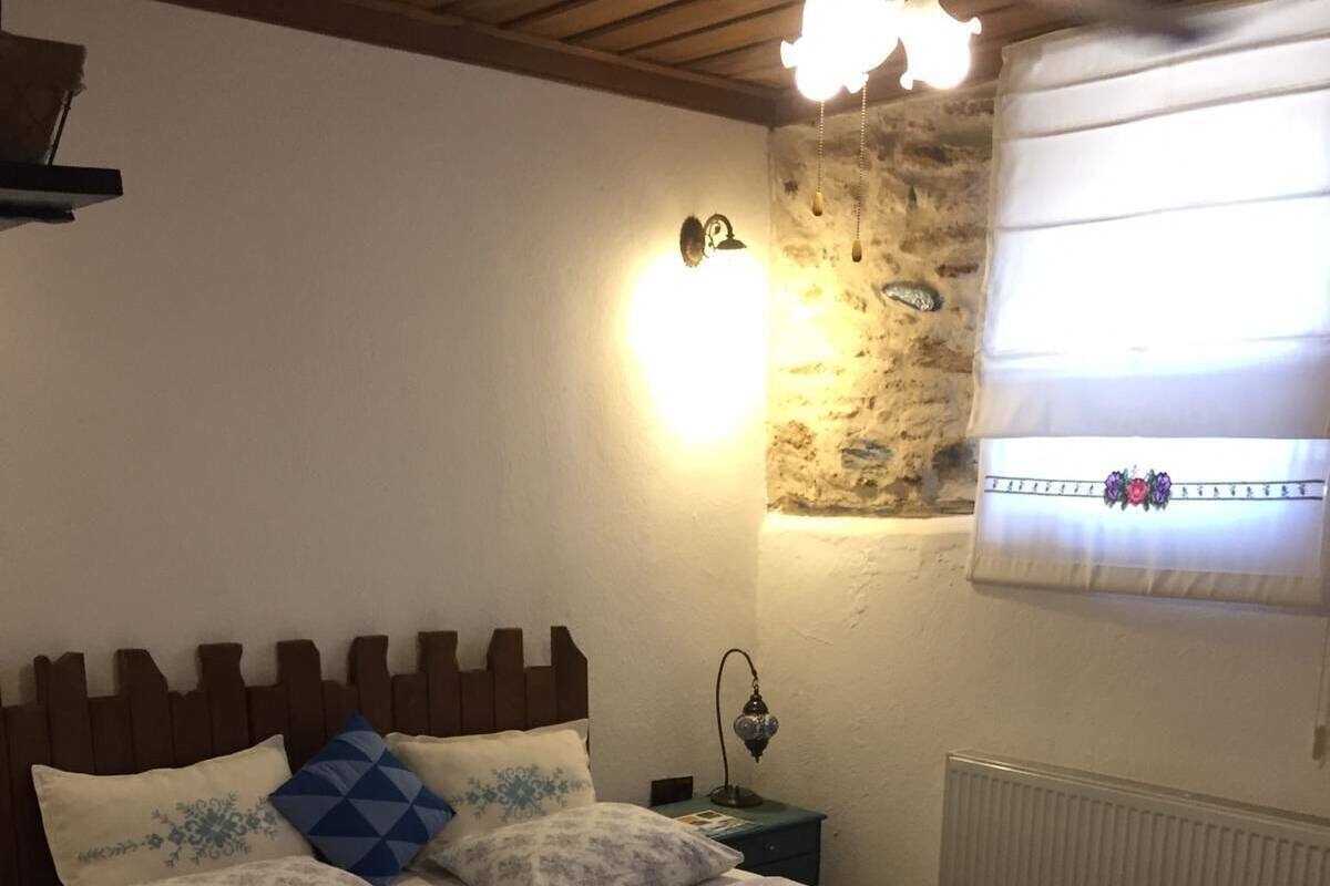 Un dormitorio con cabecera de madera, una mesita de noche con lámpara, pared de piedra y una ventana con persianas.