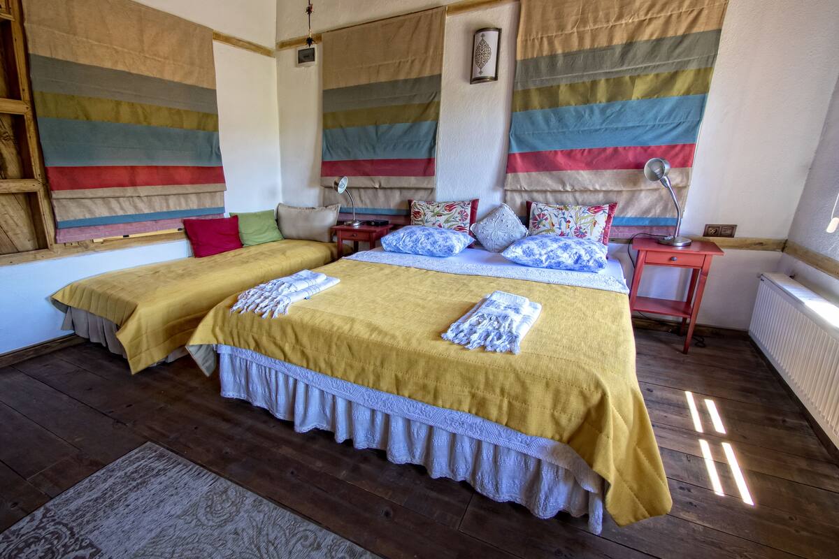 Habitación doble con dos camas, cada una con cubrecama amarillo y encaje blanco, suelo de madera y ventana con cortinas a rayas.