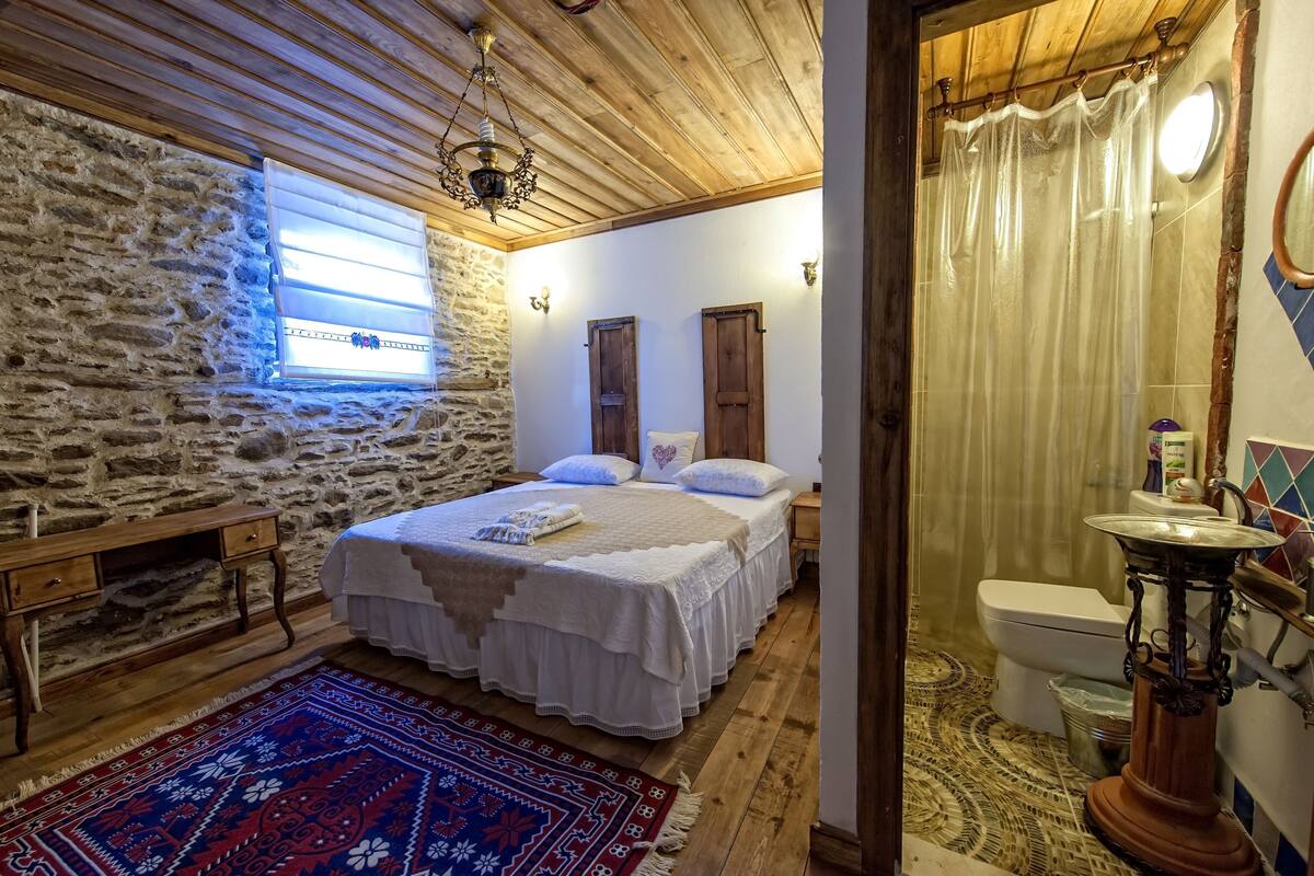 Un dormitorio con pared de piedra, techo de madera, una cama con ropa blanca, una mesita de noche, un banco de madera, una alfombra estampada, una ventana con persianas, una cortina de ducha, un inodoro y un lavabo con una base decorativa.