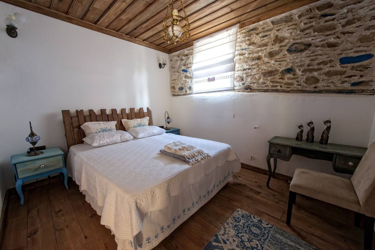 Un dormitorio con techo de madera, pared de piedra, cama con ropa de cama blanca, mesita de noche, escritorio y silla.