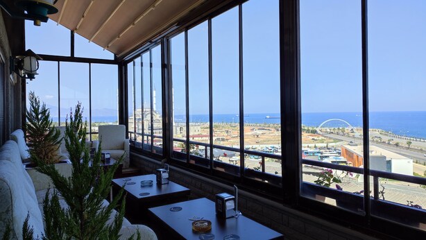 A sunlit room with large windows, a view of the sea, and outdoor seating with tables and chairs. - Funda Hotel (Trabzon)