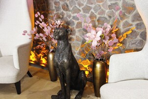 Une statue de chien est assise entre deux vases ornés de fleurs roses, dans une pièce dont le mur est en pierre et qui possède une cheminée.