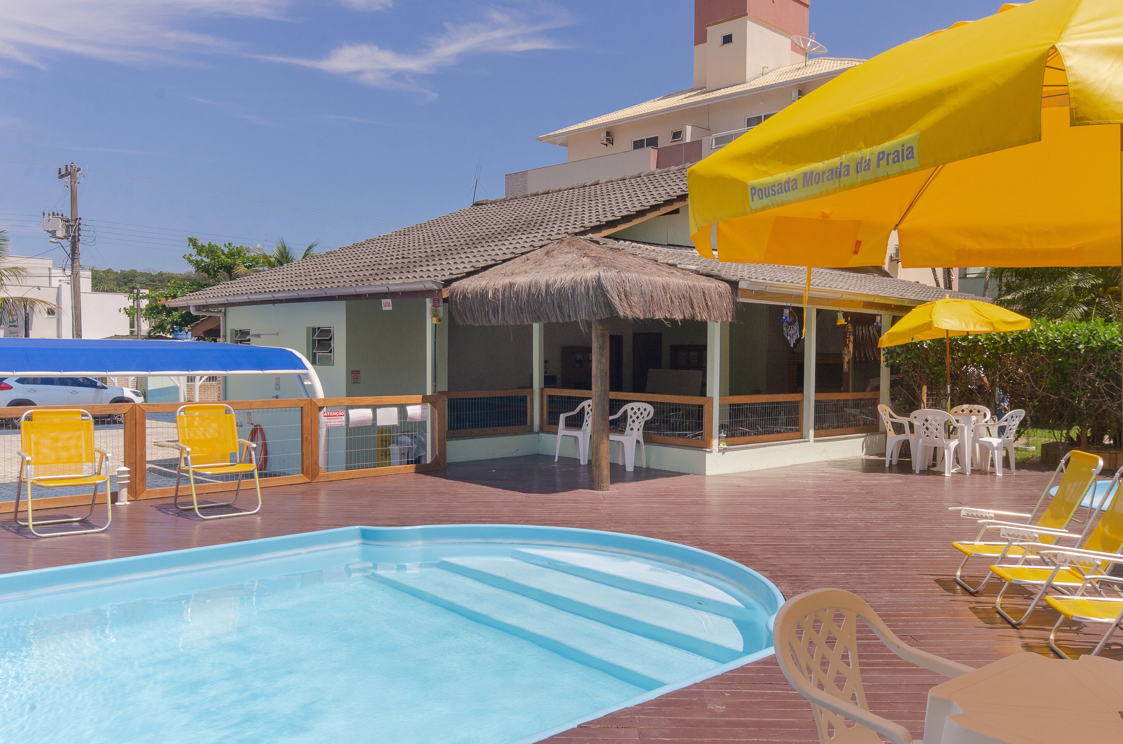 Un espace piscine agrémenté d’une structure avec toit de chaume, d’un coin salon couvert avec des parasols, et offrant une vue sur un bâtiment portant une enseigne indiquant « Pousada Morada da Praia ».
