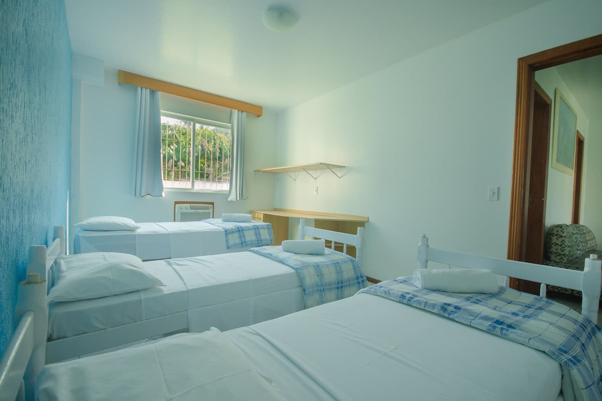 Ein Zimmer mit drei Betten, einem Fenster mit Vorhängen, einem Holzregal und einem Spiegel.