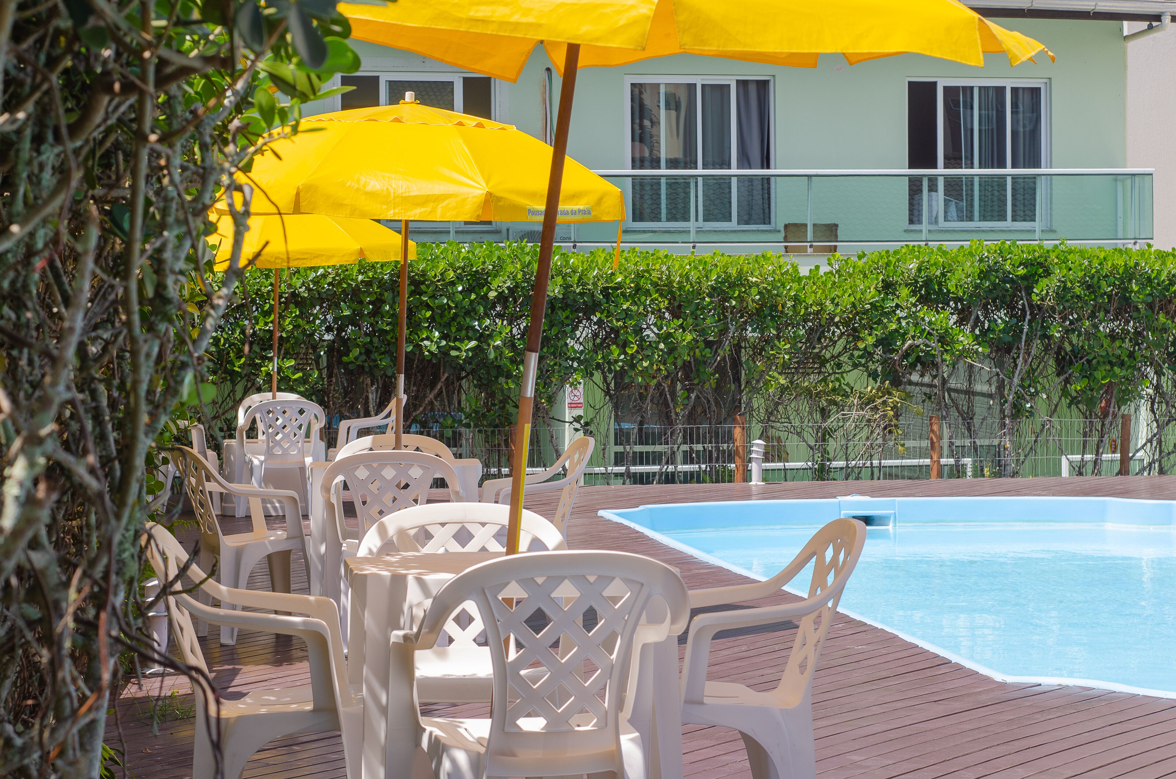 A poolside area with white plastic chairs and yellow umbrellas, surrounded by greenery and a building in the background.