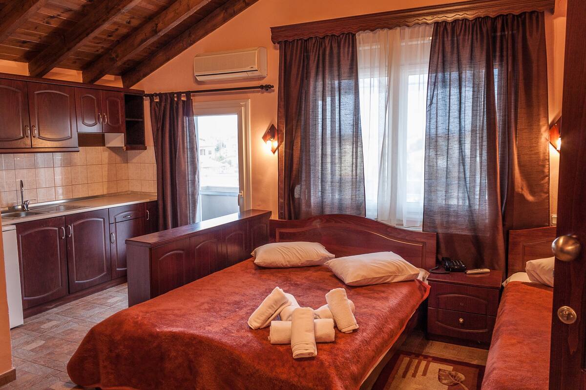 Ein Schlafzimmer mit einem großen Bett, einem Nachttisch, Vorhängen an einem Fenster sowie einer Küchenzeile mit Schränken und einem Spülbecken.