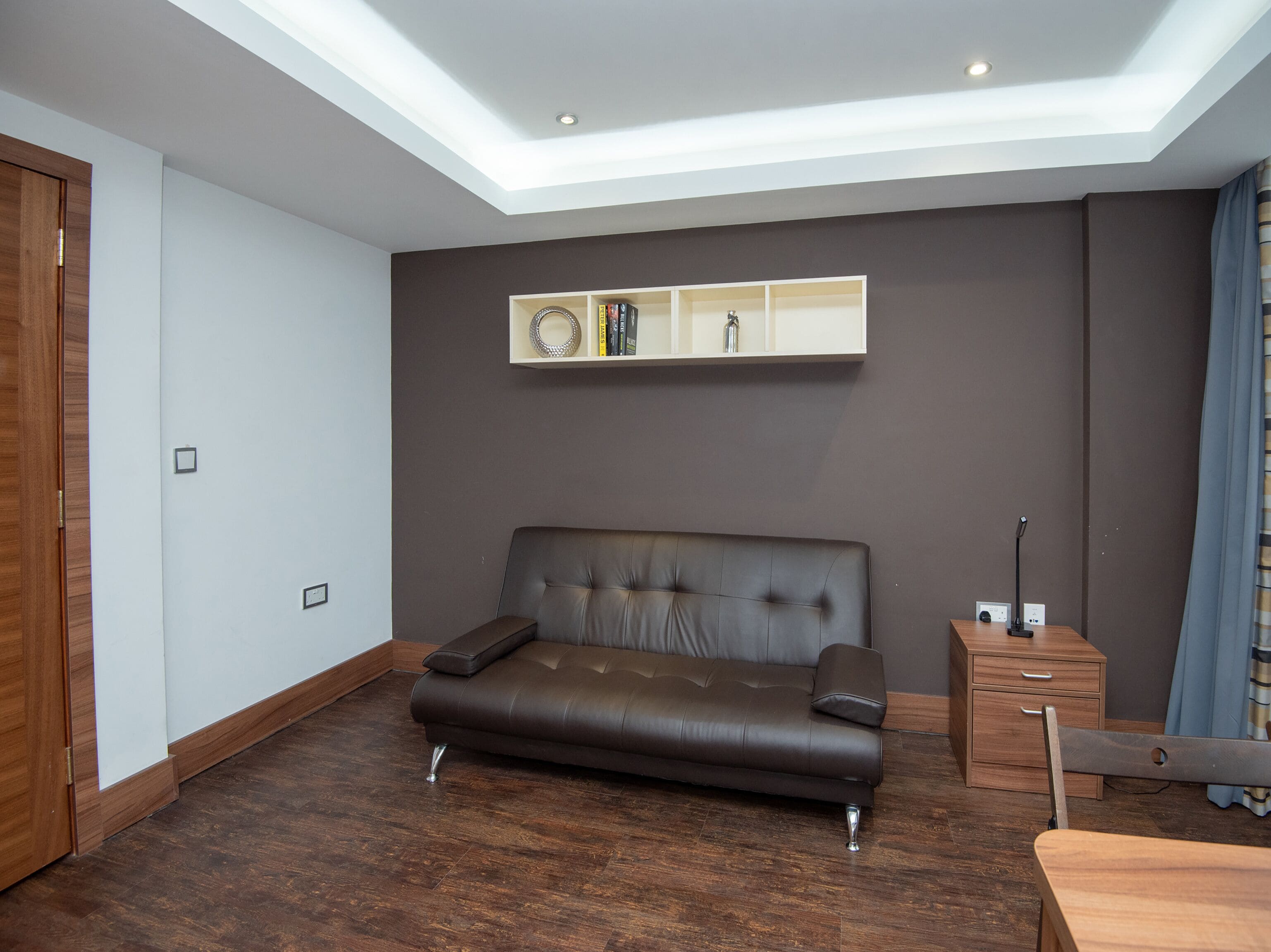 Un salon moderne avec un canapé en cuir marron foncé, une table d’appoint en bois et une étagère intégrée où sont disposés des livres et des objets décoratifs.