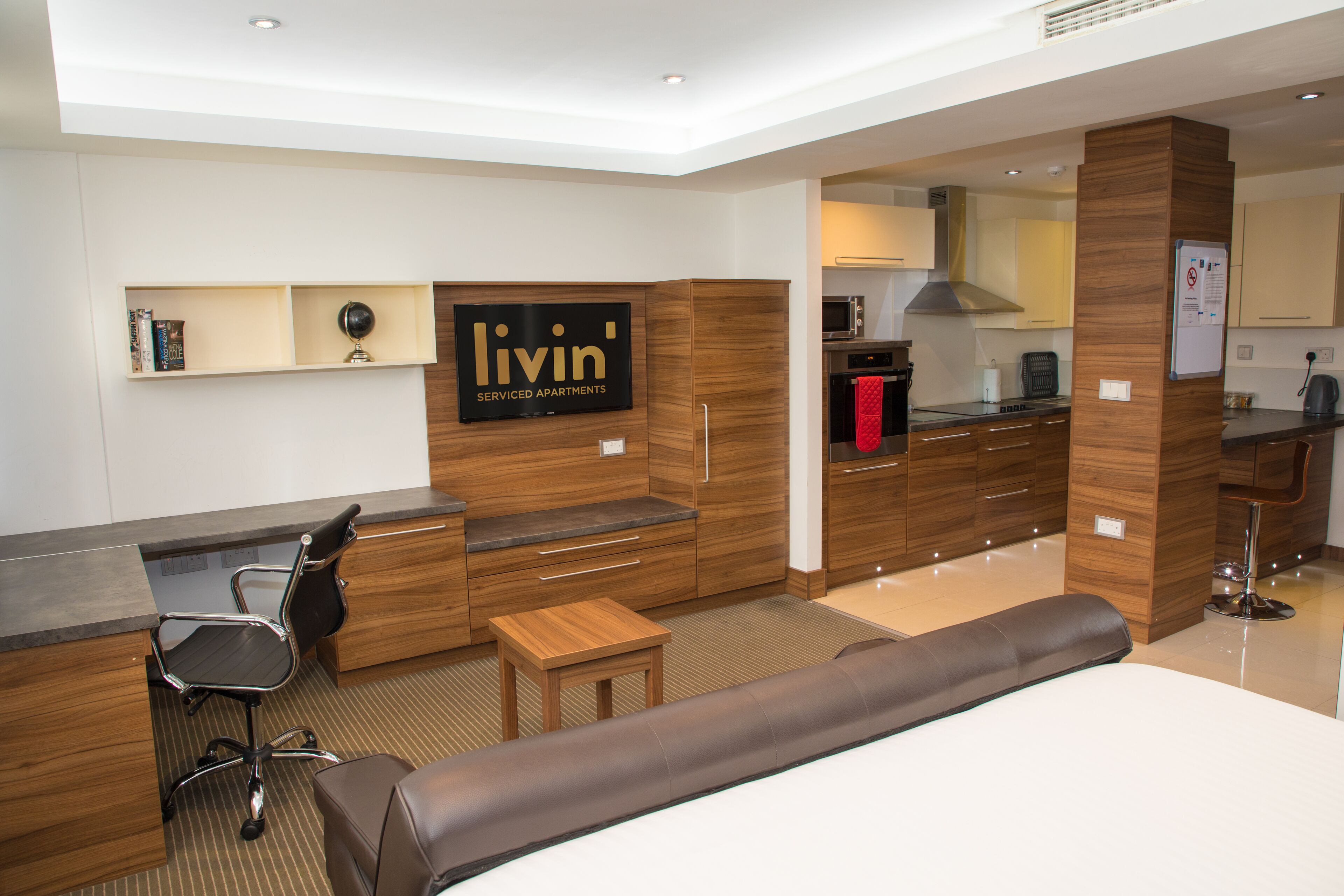 A modern hotel room with a bed, a desk, a chair, a small table, a sofa, a kitchen area, and a mounted TV.