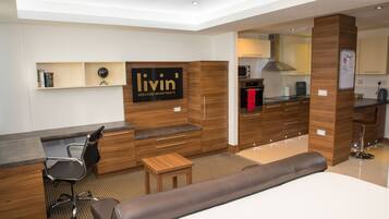 A modern hotel room with a bed, a desk, a chair, a small table, a sofa, a kitchen area, and a mounted TV.