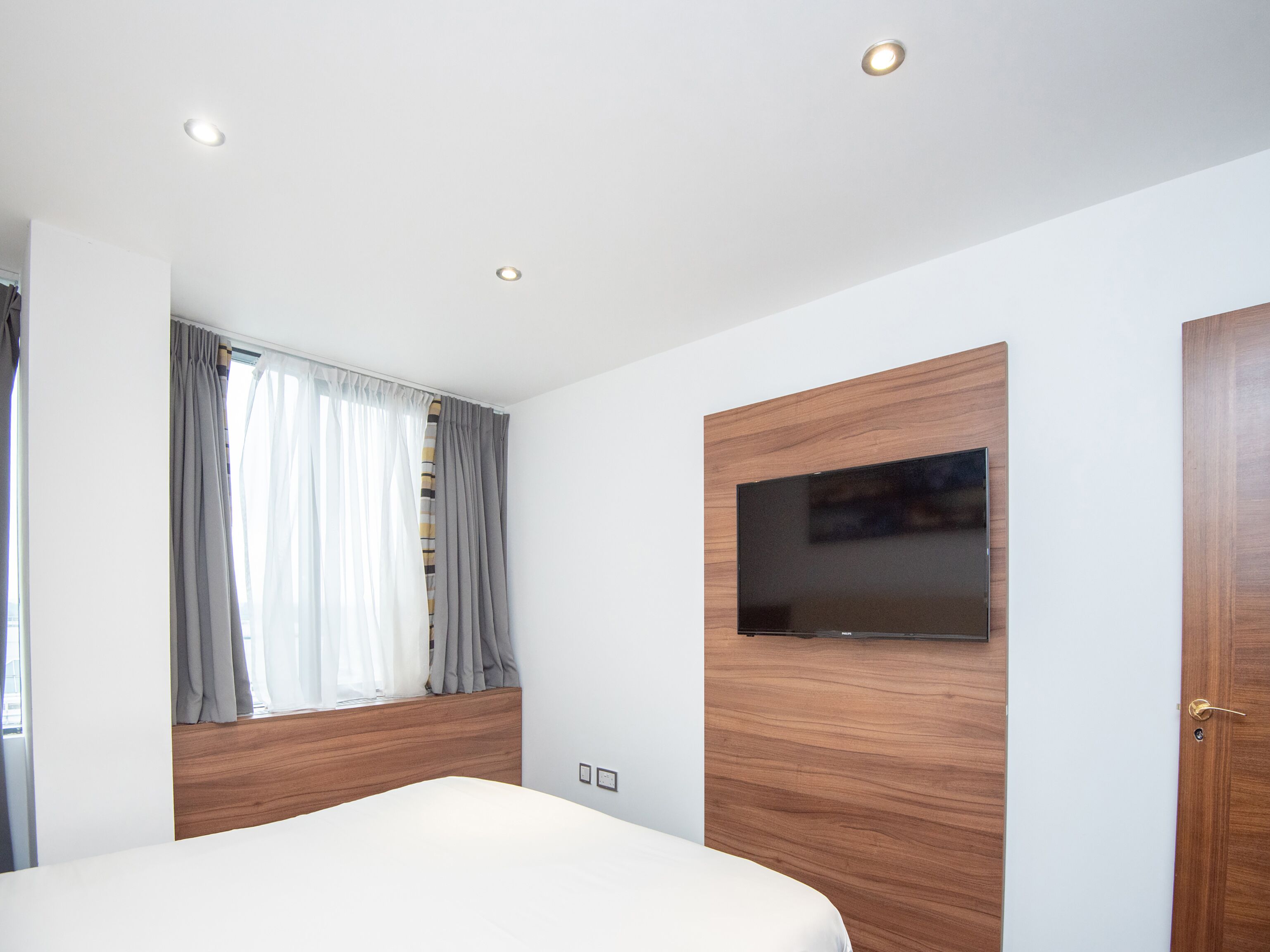 A modern bedroom with a bed, a TV mounted on a wooden panel, a window with curtains, and recessed ceiling lights.