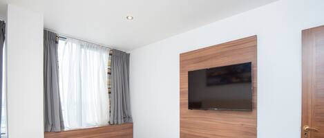 A modern bedroom with a bed, a TV mounted on a wooden panel, a window with curtains, and recessed ceiling lights.
