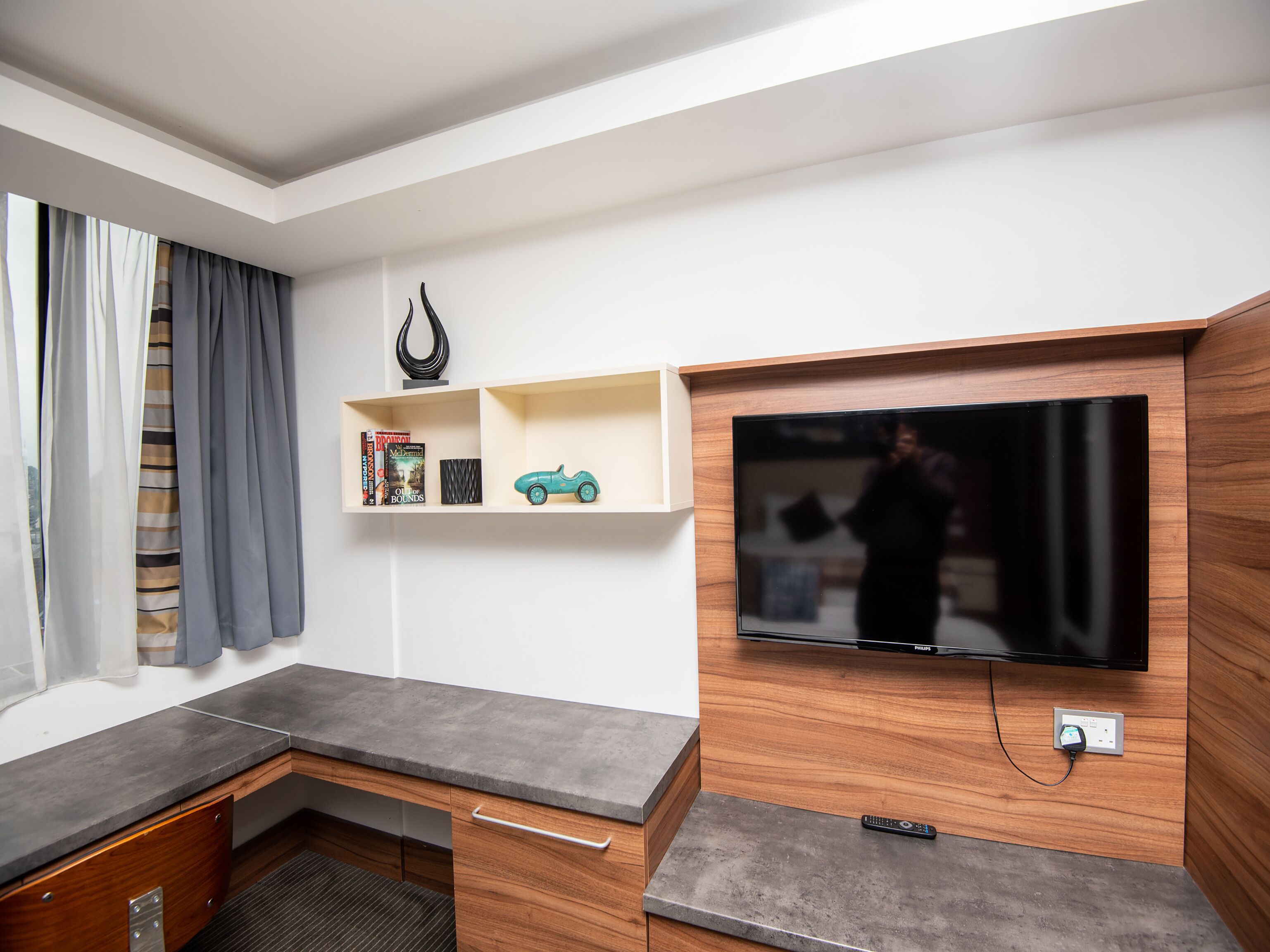 A modern living room with a flat-screen TV mounted on a wooden wall, a grey countertop, and a small wooden shelf with books and a toy car.
