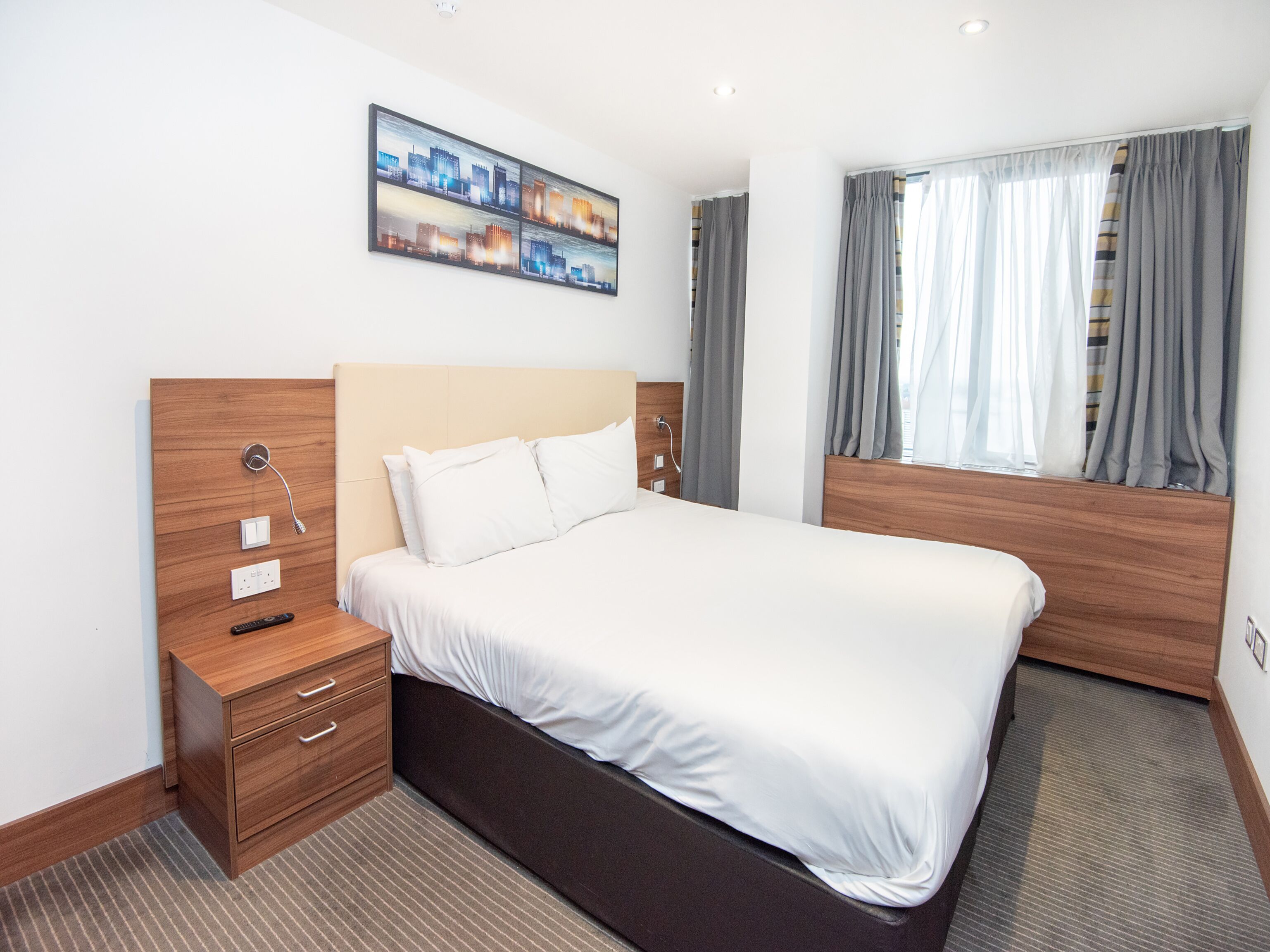 A hotel room with a large bed, wooden headboard, a nightstand with drawers, a bedside table with a lamp, a painting on the wall, and a window with curtains.