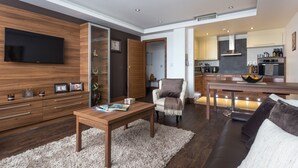A modern living room with a wooden TV stand, a coffee table, and a sofa. There is a kitchen visible in the background with a dining area. - Livin’ Serviced Apartments (Watford)