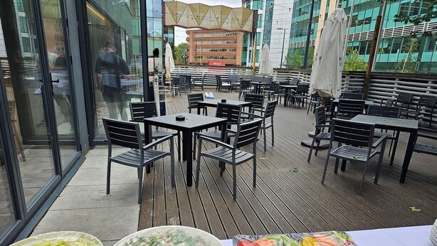 An outdoor dining area with tables and chairs, surrounded by glass walls and a view of buildings. - Livin’ Serviced Apartments (Watford)