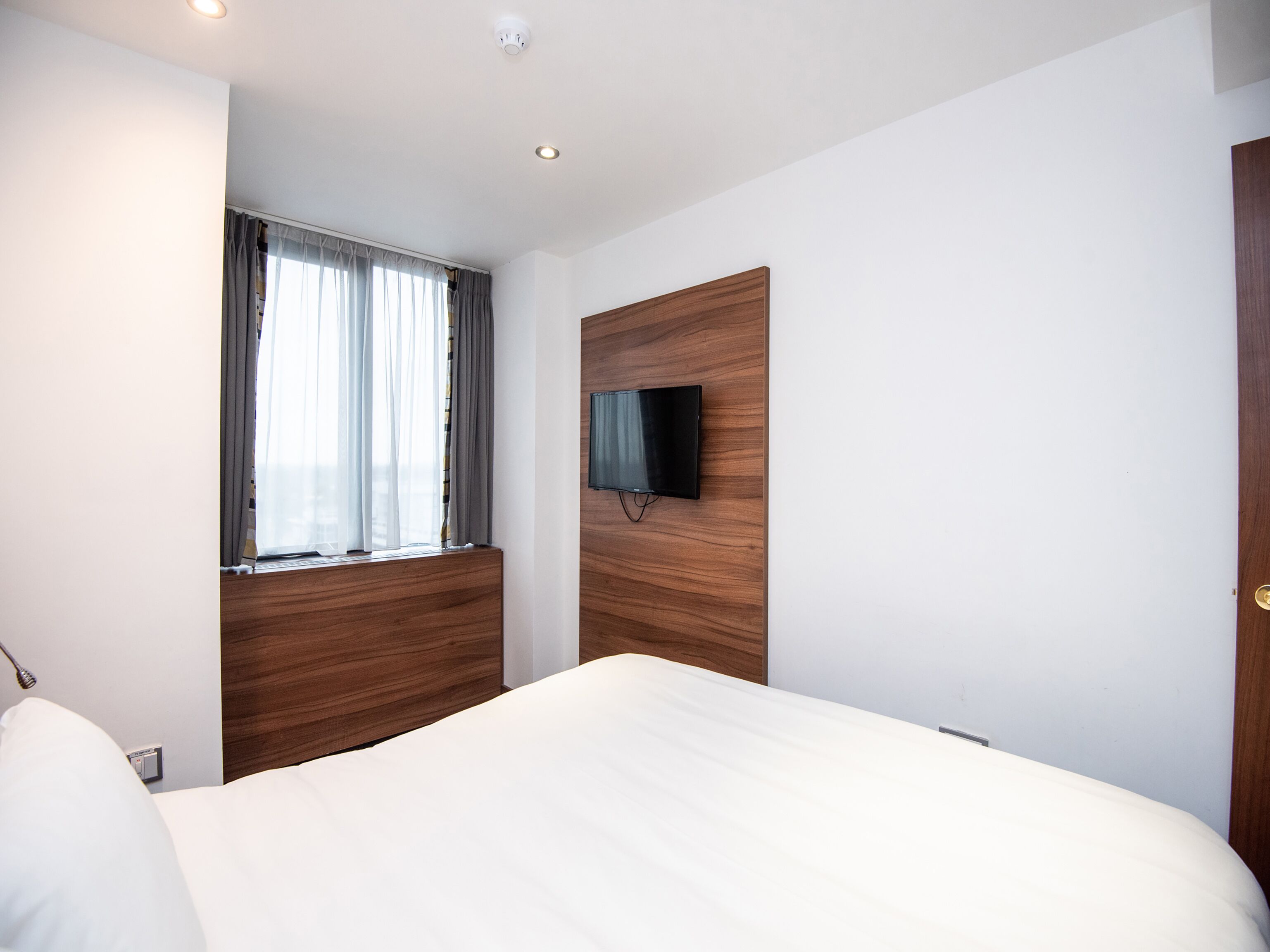 Una habitación de hotel moderna con una cama grande, un televisor de pantalla plana montado en un panel de madera, una ventana con cortinas translúcidas y una lámpara en el techo.