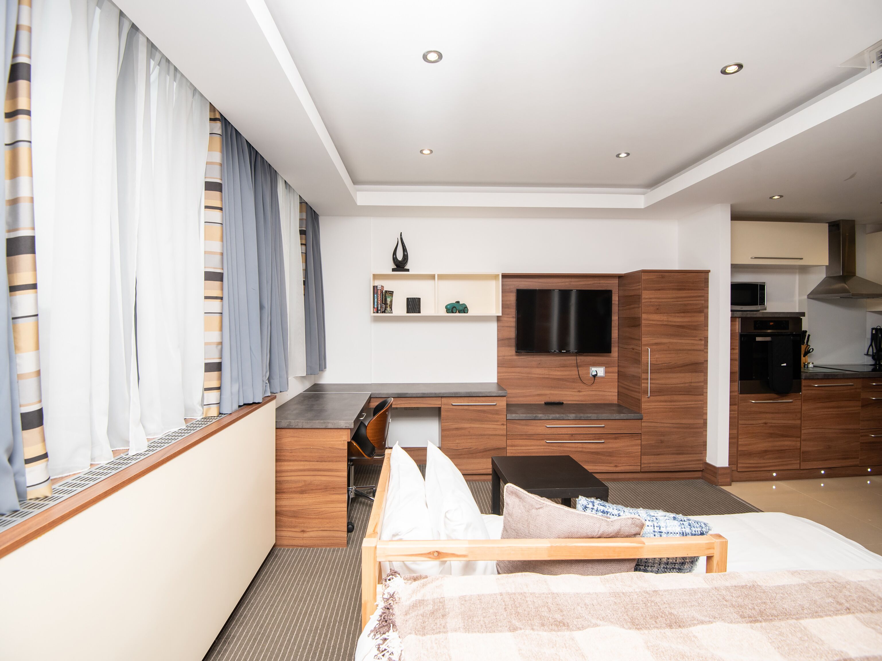 A modern hotel room with a bed, a desk, a TV, and a kitchenette.