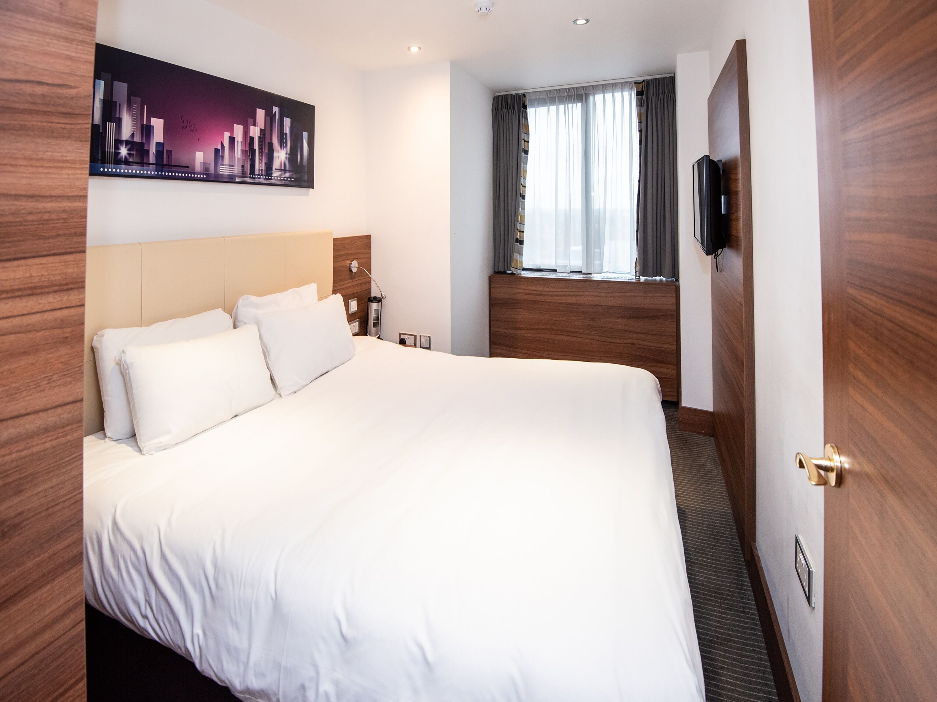 A hotel room with a large bed, a headboard, a nightstand, a lamp, a window with curtains, and a wall-mounted TV.