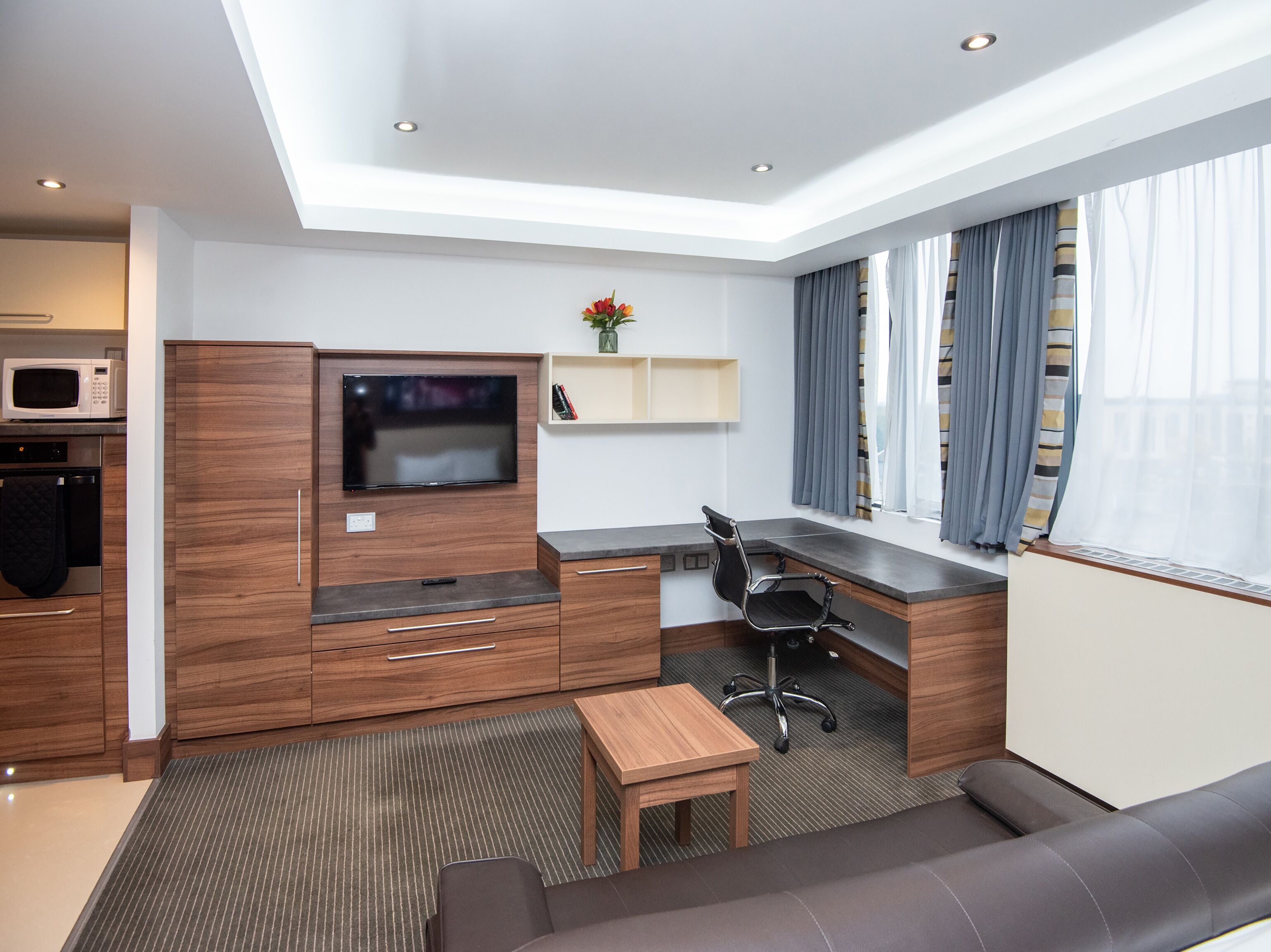 Habitació d'hotel moderna amb cuina americana, televisió de pantalla plana penjada en un moble de fusta, escriptori amb cadira, sofà i una finestra gran amb cortines.