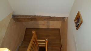 Un escalier en bois avec une rampe, un plafond avec des poutres apparentes et un tableau encadré au mur.