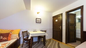 Une petite pièce agréable et confortable, avec un lit, une table et des chaises. Un tableau encadré est accroché au mur, et une porte donne accès à une autre pièce.