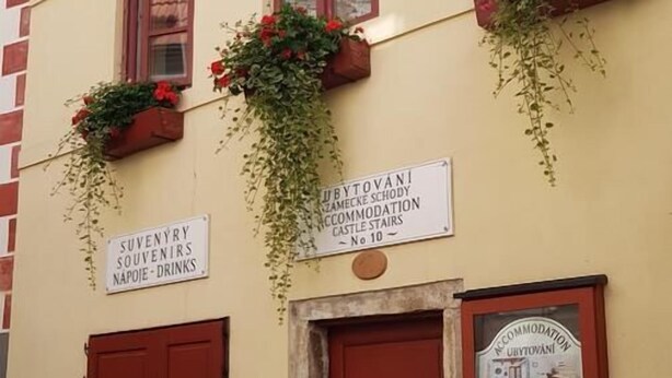 Un bâtiment avec une porte rouge, deux fenêtres avec des volets rouges et des jardinières sur les rebords de fenêtre. On y voit des panneaux indiquant « SOUVENIRS » et « HÉBERGEMENT ».