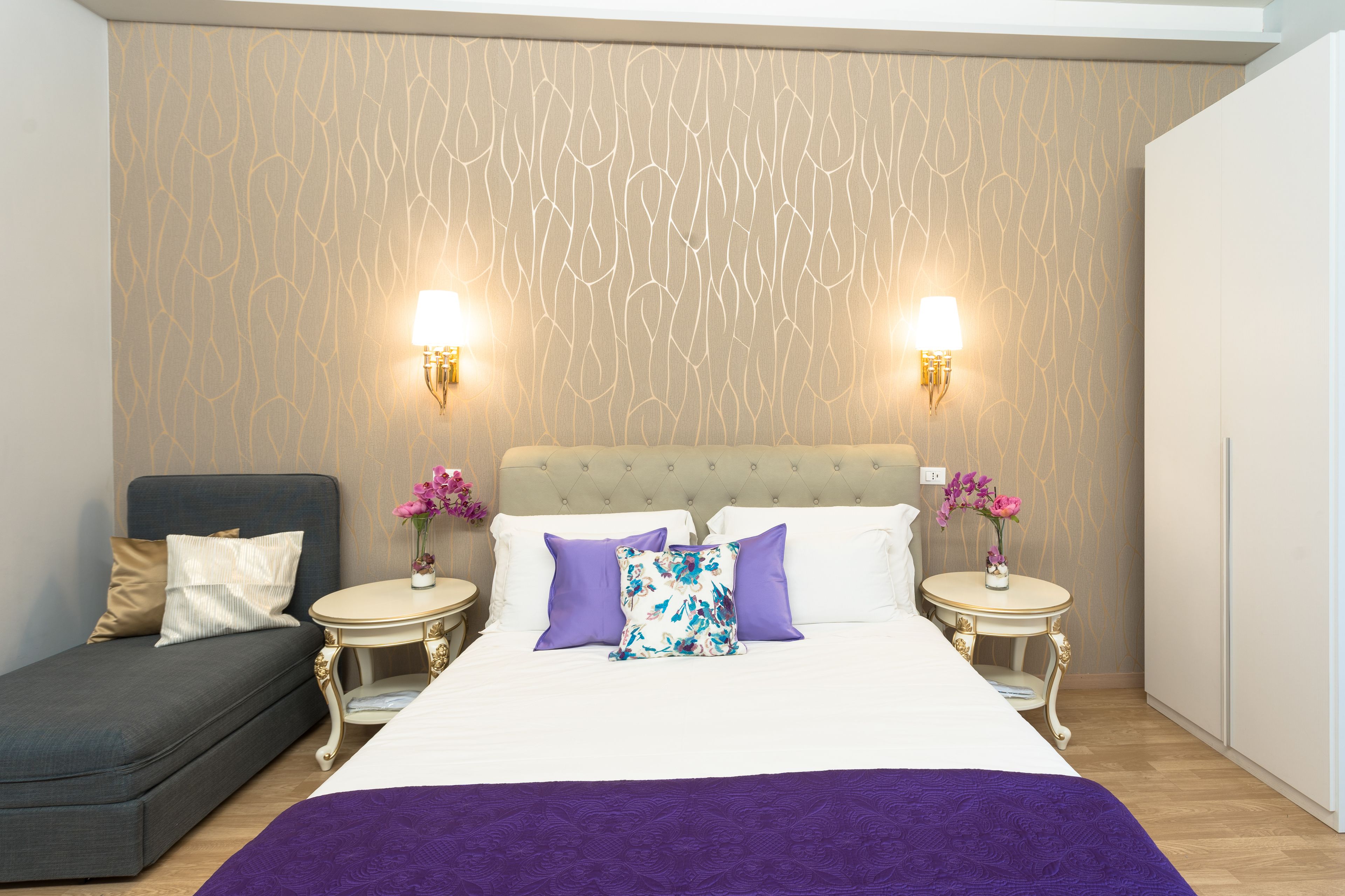 A neatly arranged bedroom with a bed, a sofa, two nightstands with lamps, and decorative flowers.