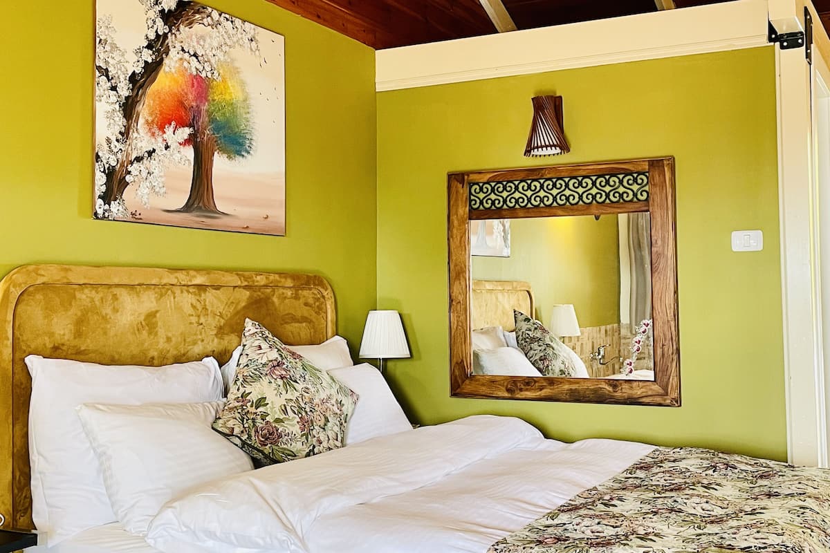 A bed with white linens and floral pillows, a wooden headboard, a mirror with a decorative frame, and a painting of a tree on the wall.