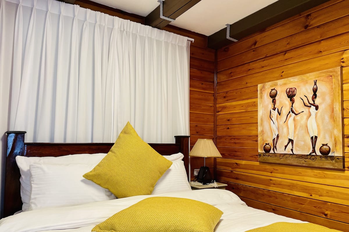 A hotel room with a wooden paneled wall, a bed with white bedding and yellow pillows, a painting of figures holding pots, and a window with white curtains.