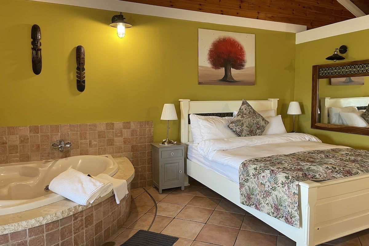 A bedroom with a bed, bedside lamps, a nightstand, a painting, and a bathroom with a bathtub and towel.