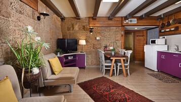 Un salon moderne avec un mur en pierre, des poutres en bois, un téléviseur, un canapé, une table à manger et un coin cuisine doté de meubles violets.