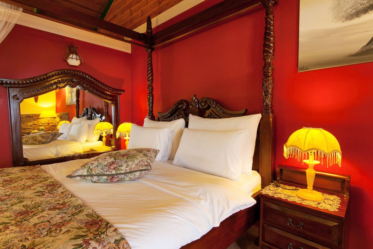 A bedroom with a four-poster bed, a nightstand with a lamp, and a mirror above the bed.